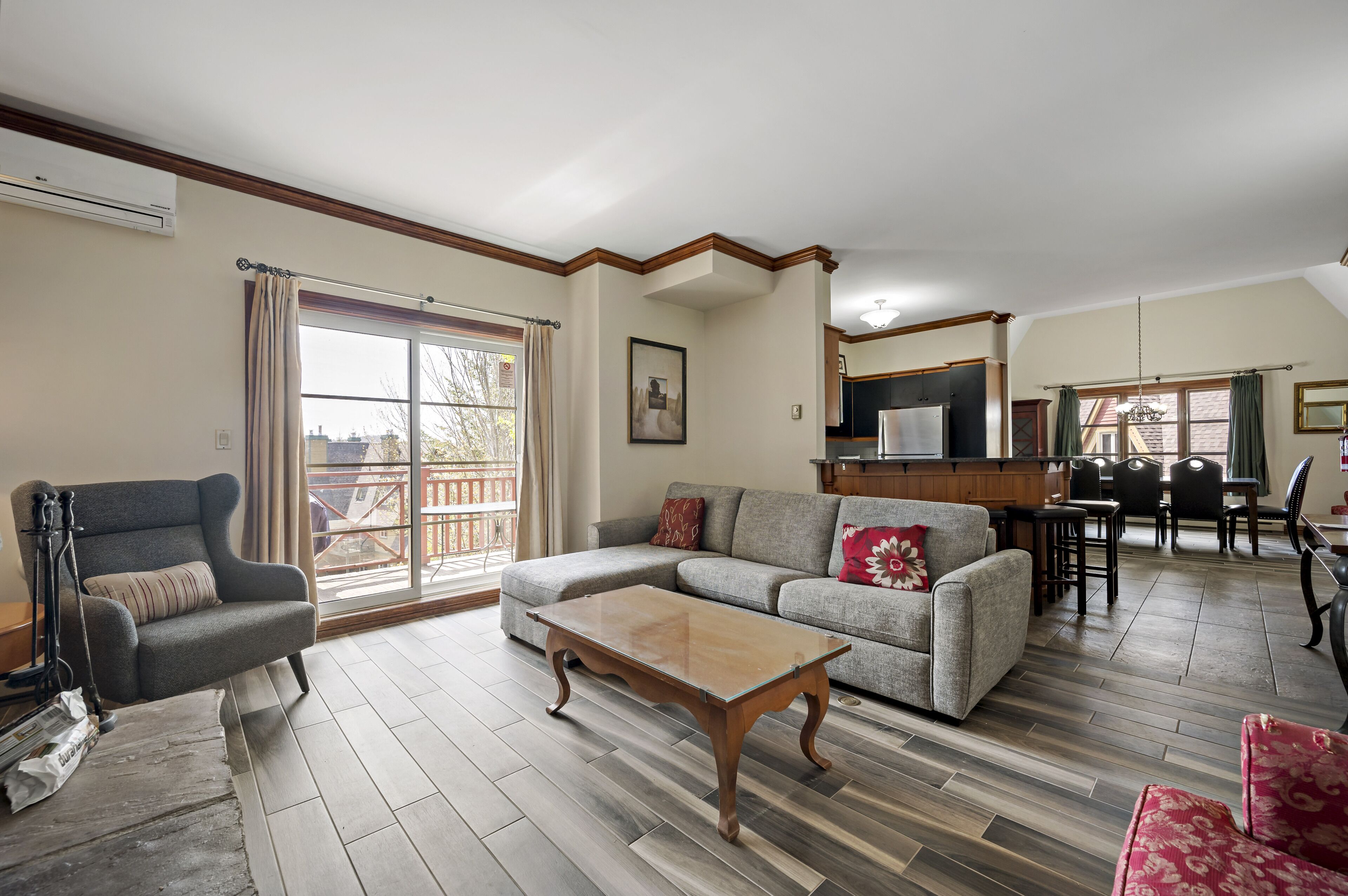 A living room with a grey sofa, a wooden coffee table, and a grey armchair. There is a dining area with a table and chairs, and a kitchen area with a countertop and cabinets.