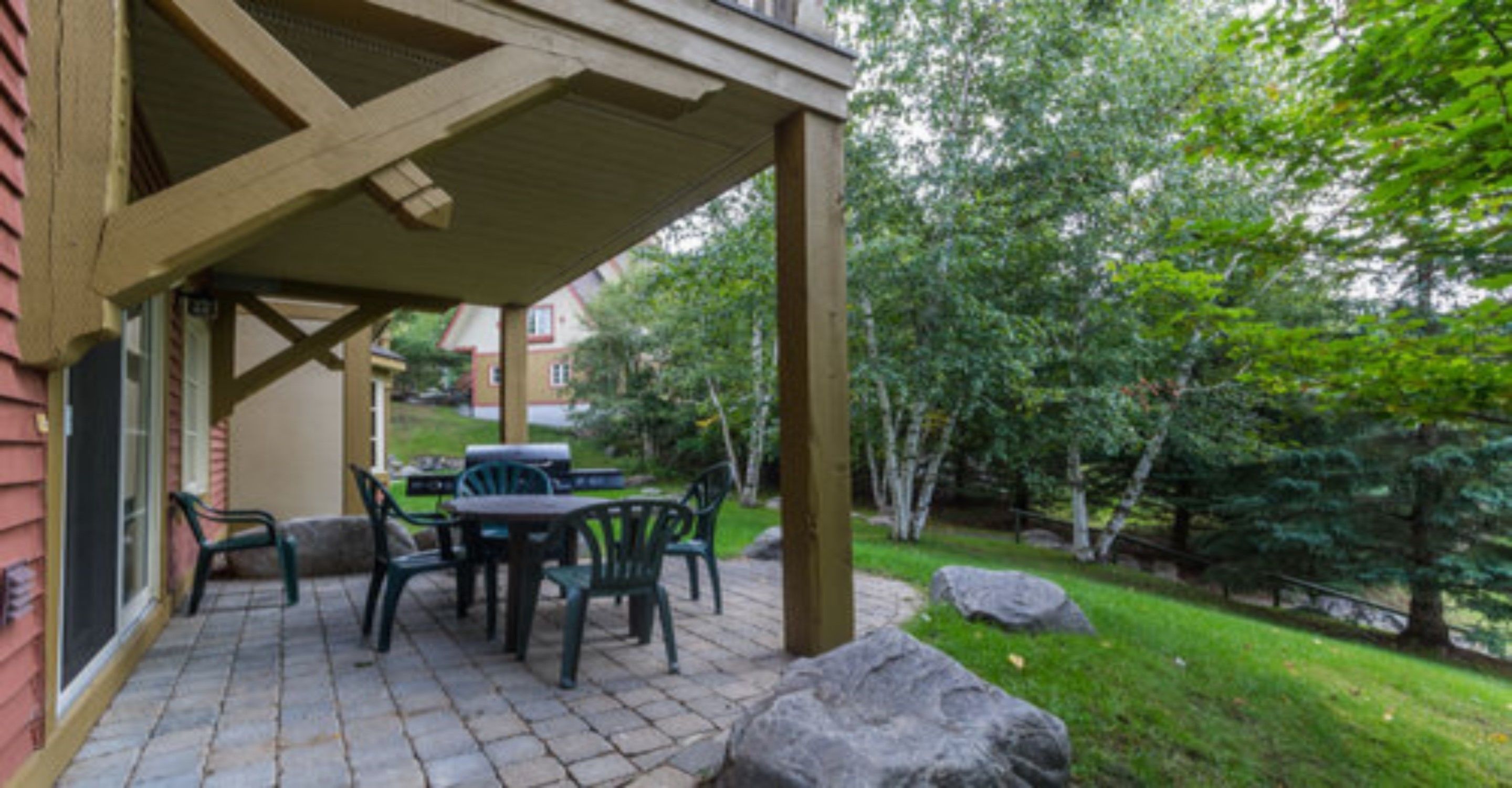 Une terrasse couverte avec un sol en briques, des meubles d’extérieur et une vue sur un quartier résidentiel, avec des arbres et une maison en arrière-plan.