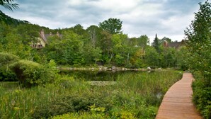 A wooden boardwalk leading through a lush garden with a pond and dense greenery. - Le Plateau - RVMT (Mont-Tremblant)
