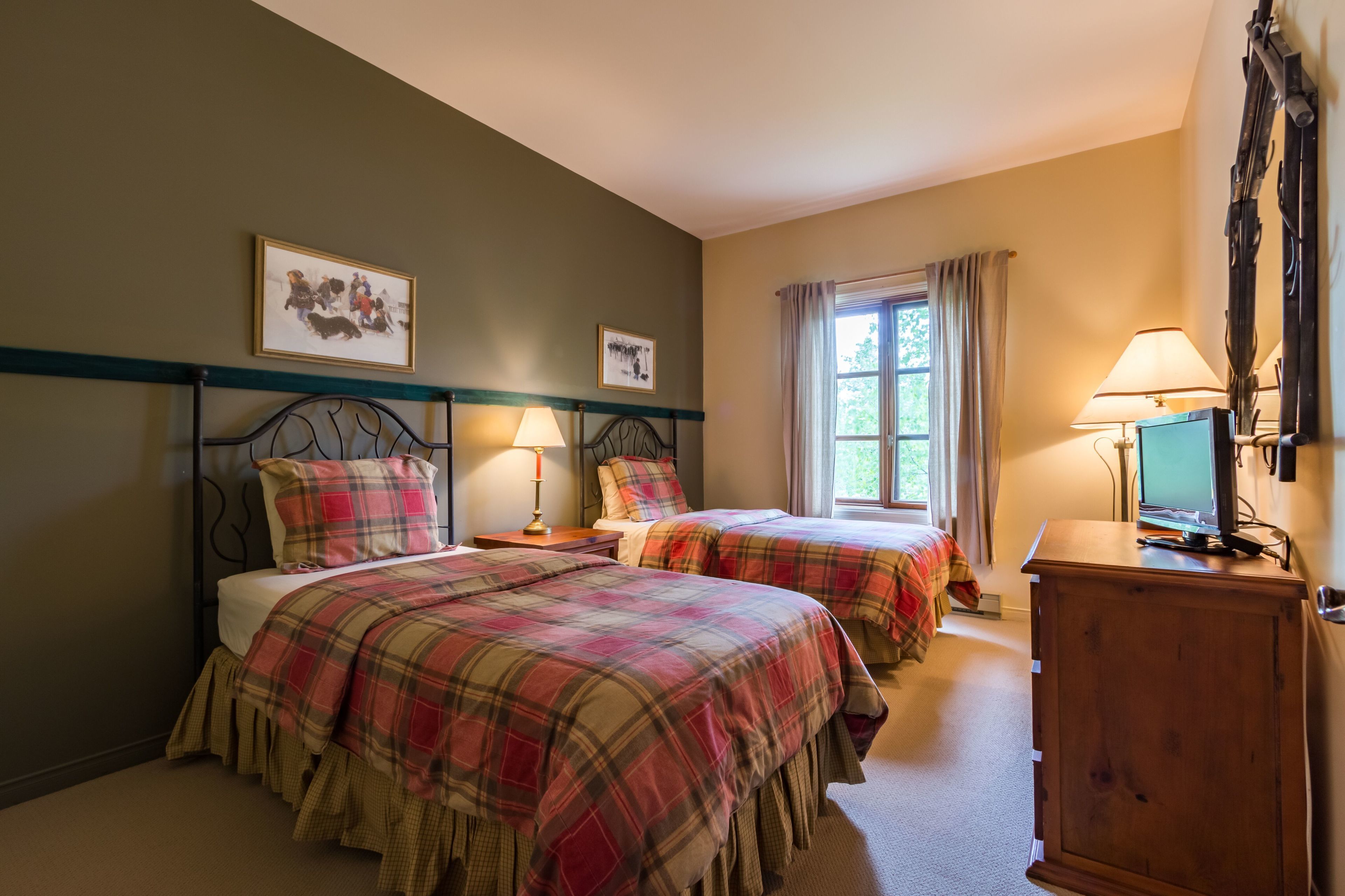 A room with two beds, each with a plaid bedspread, a wooden cabinet, and a window with curtains.