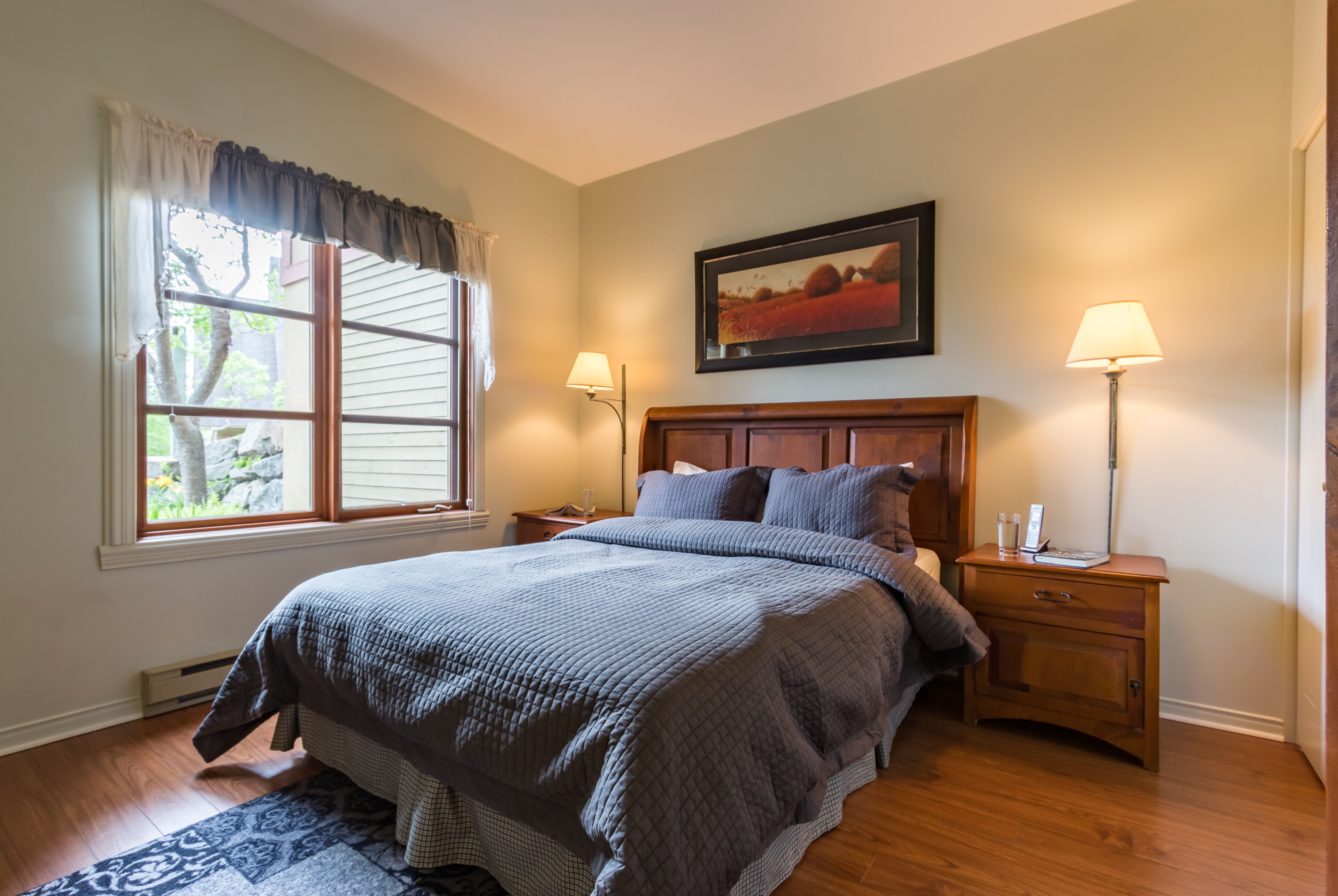 A bedroom with a large bed, two bedside tables, a nightstand, a window with curtains, a painting on the wall, and a lamp on each bedside table.