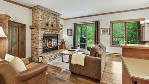A living room with a stone fireplace, wooden furniture, and large windows with a view of greenery. - Le Plateau - RVMT (Mont-Tremblant)