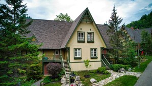 A traditional house with a steeply pitched roof, a stone pathway leading to the front door, and a small garden with rocks and shrubs. - Le Plateau - RVMT (Mont-Tremblant)