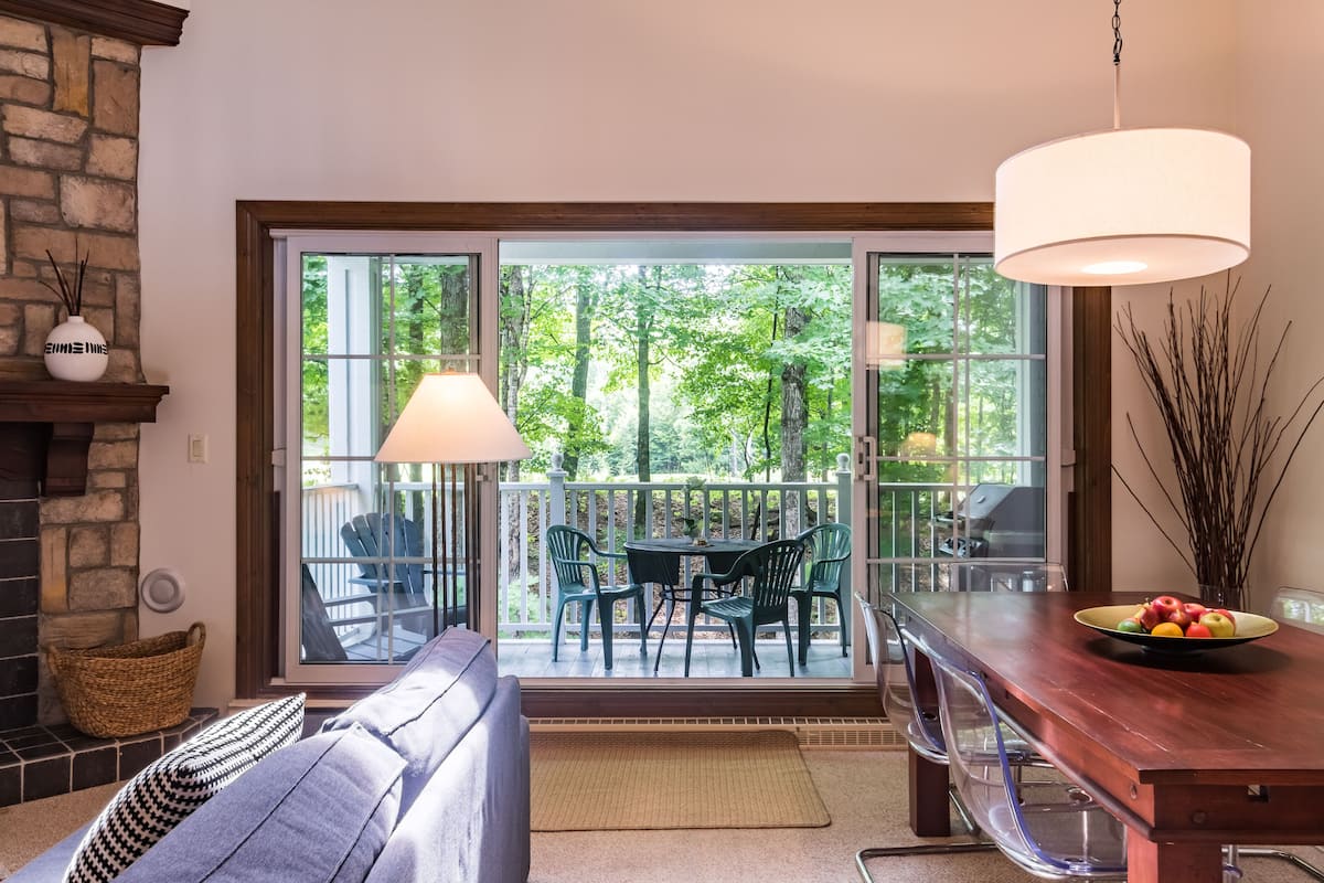 A modern living room with a fireplace, a sofa, a dining table with chairs, and a large sliding glass door leading to a deck with a view of trees.