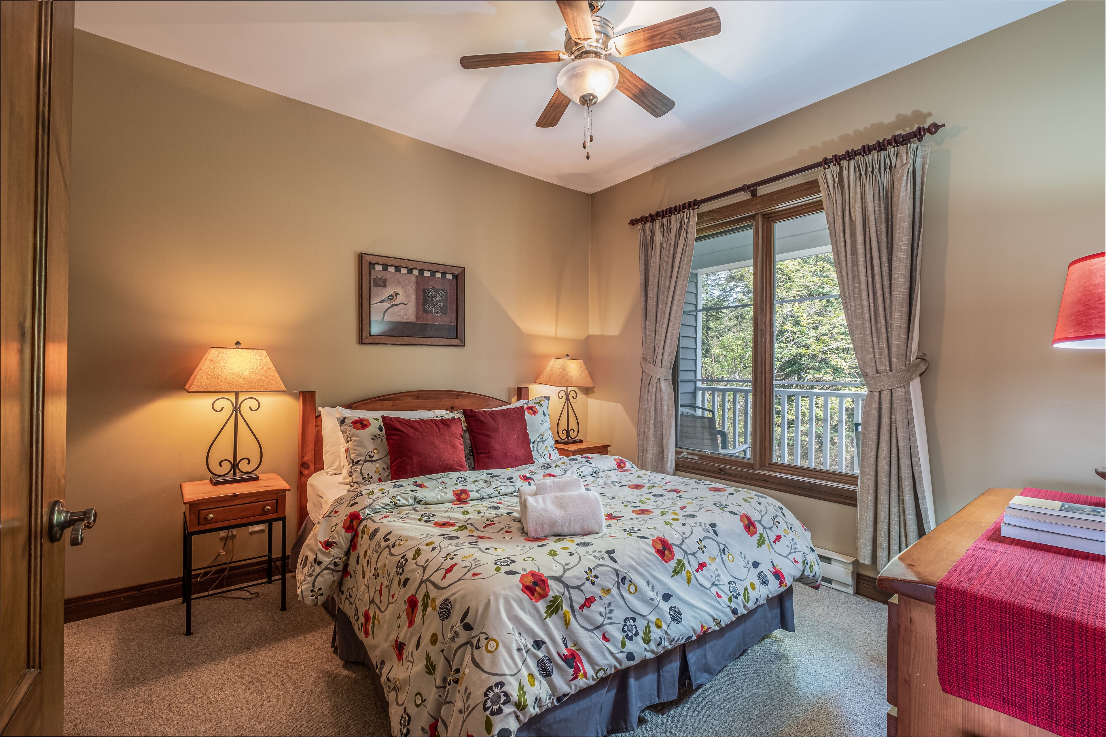 A bedroom with a bed, two bedside lamps, a ceiling fan, a window with curtains, a framed picture on the wall, and a small wooden table with a red cloth.