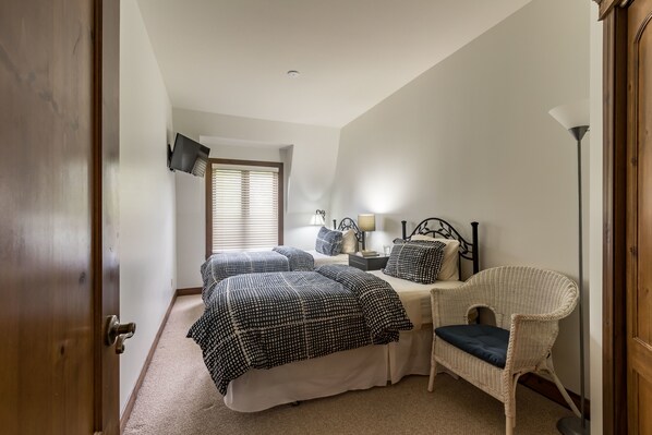 A bedroom with a bed, bedside table, lamp, chair, and a window with blinds. - Les Manoirs - RVMT (Mont-Tremblant)