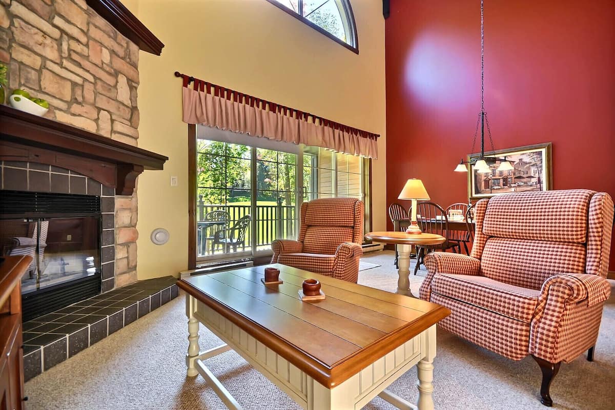 A cozy living room with a fireplace, checkered armchairs, a wooden coffee table, and a dining table with chairs.