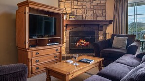 A cozy living room with a stone fireplace, a wooden entertainment unit, and comfortable seating. - Les Manoirs - RVMT (Mont-Tremblant)