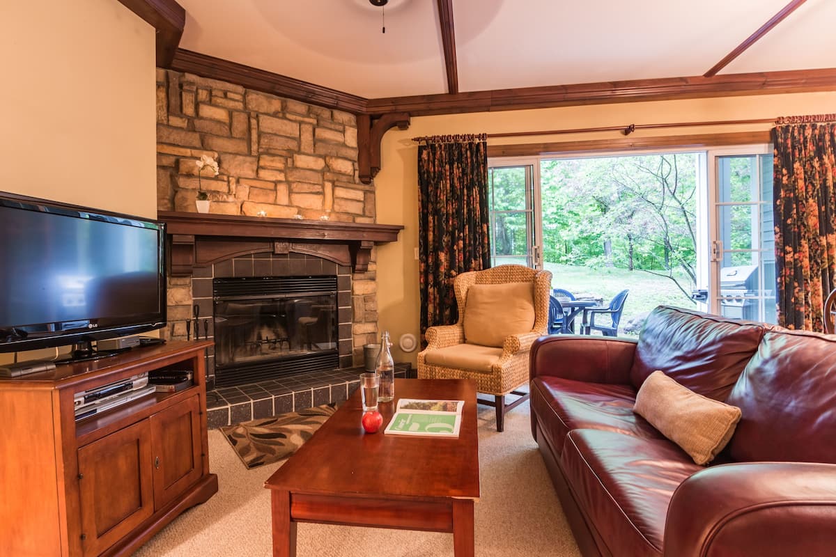 A cozy living room with a stone fireplace, a leather sofa, a television, and a dining area visible through the glass doors.