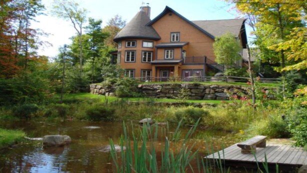 A large, two-story house with a stone foundation and a wrap-around porch, surrounded by a pond and lush greenery. - Les Manoirs - RVMT (Mont-Tremblant)