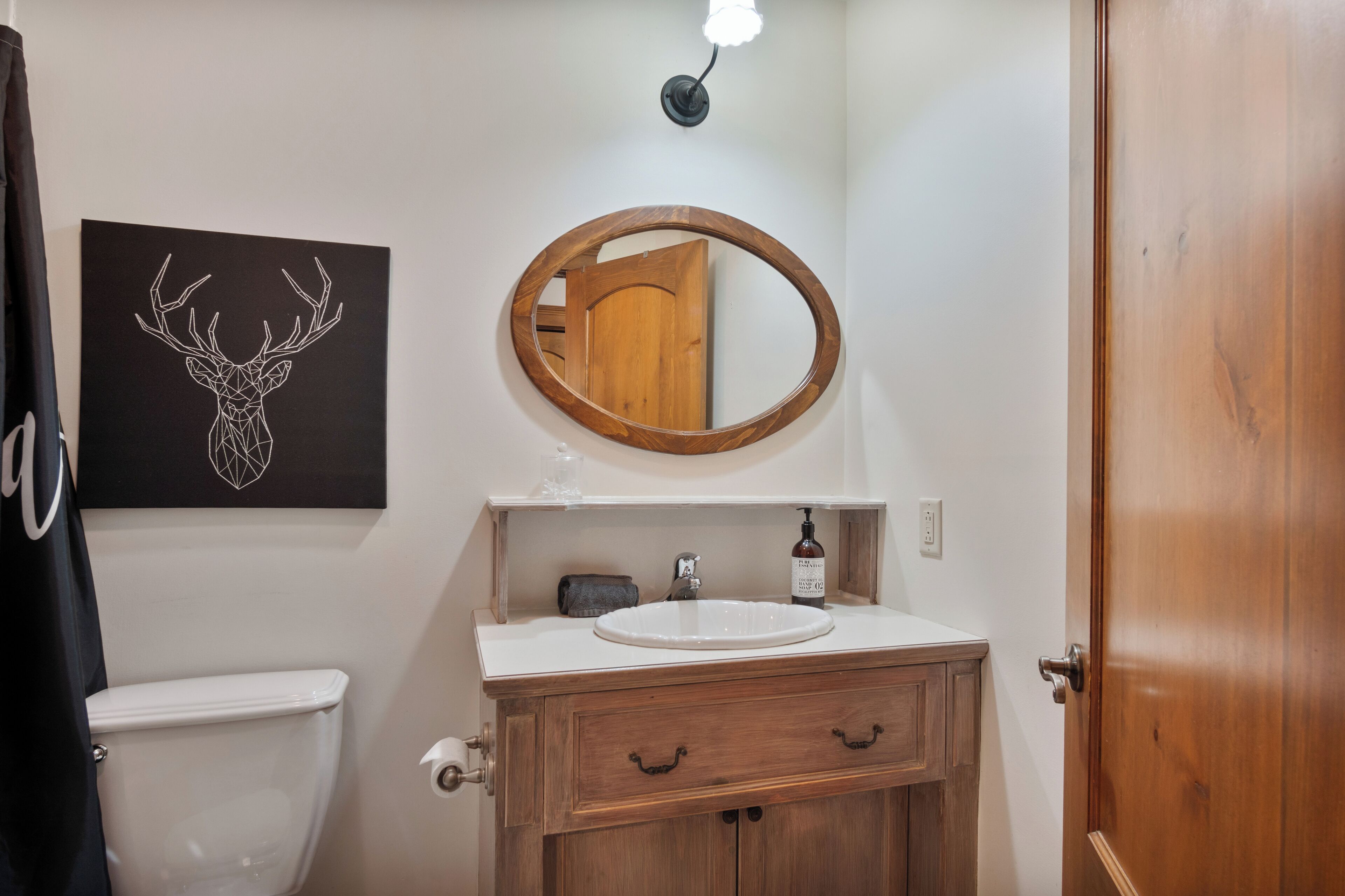 A bathroom with a wooden vanity, a round mirror, a white sink, and a deer head artwork on the wall.