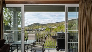 An open sliding glass door leading to a balcony with a grill, a table with chairs, and a view of a hilly landscape. - Les Manoirs - RVMT (Mont-Tremblant)