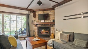 A cozy living room with a fireplace, a sofa, a coffee table, and a television mounted on a stone wall. - Les Manoirs - RVMT (Mont-Tremblant)