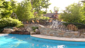 A swimming pool with a stone and metal deck, surrounded by lush greenery and a stone wall. - Les Manoirs - RVMT (Mont-Tremblant)