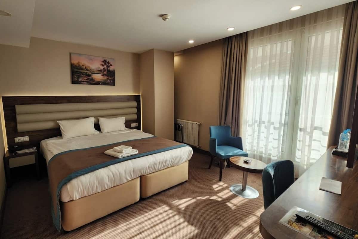 Habitación de hotel con una cama grande, un escritorio con silla, un cuadro en la pared y una ventana con cortinas.