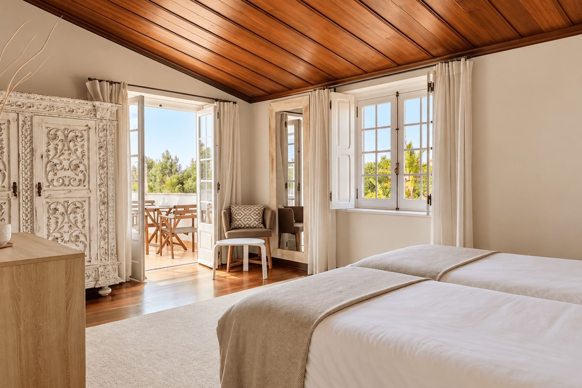 A bedroom with two beds, a wooden ceiling, a balcony with a table and chairs, and a view of greenery.
