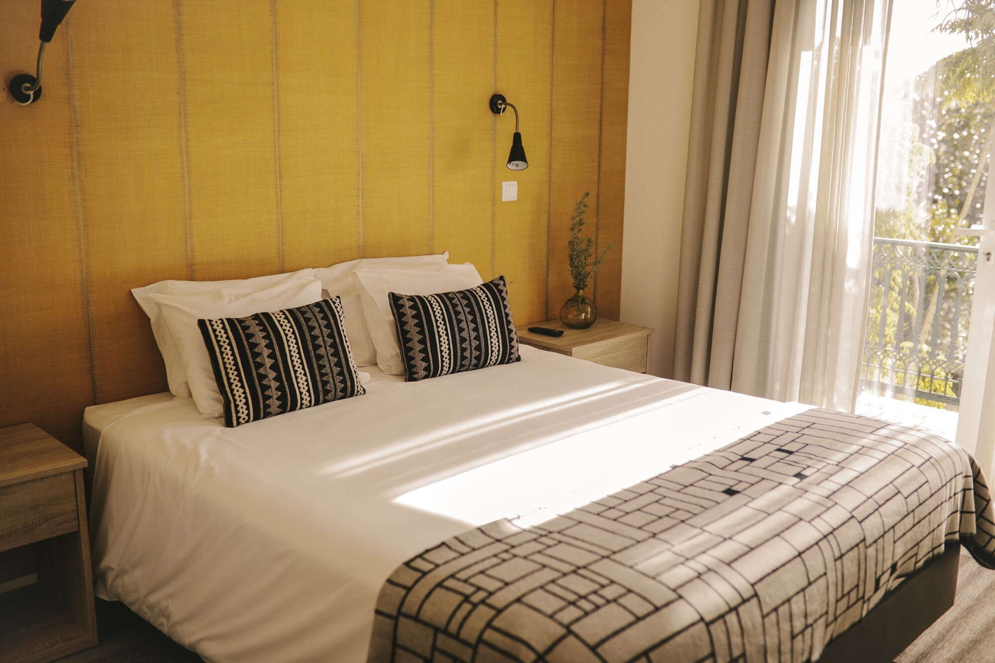 A neatly made bed with patterned pillows, a bedside lamp, and a view of greenery through sheer curtains.