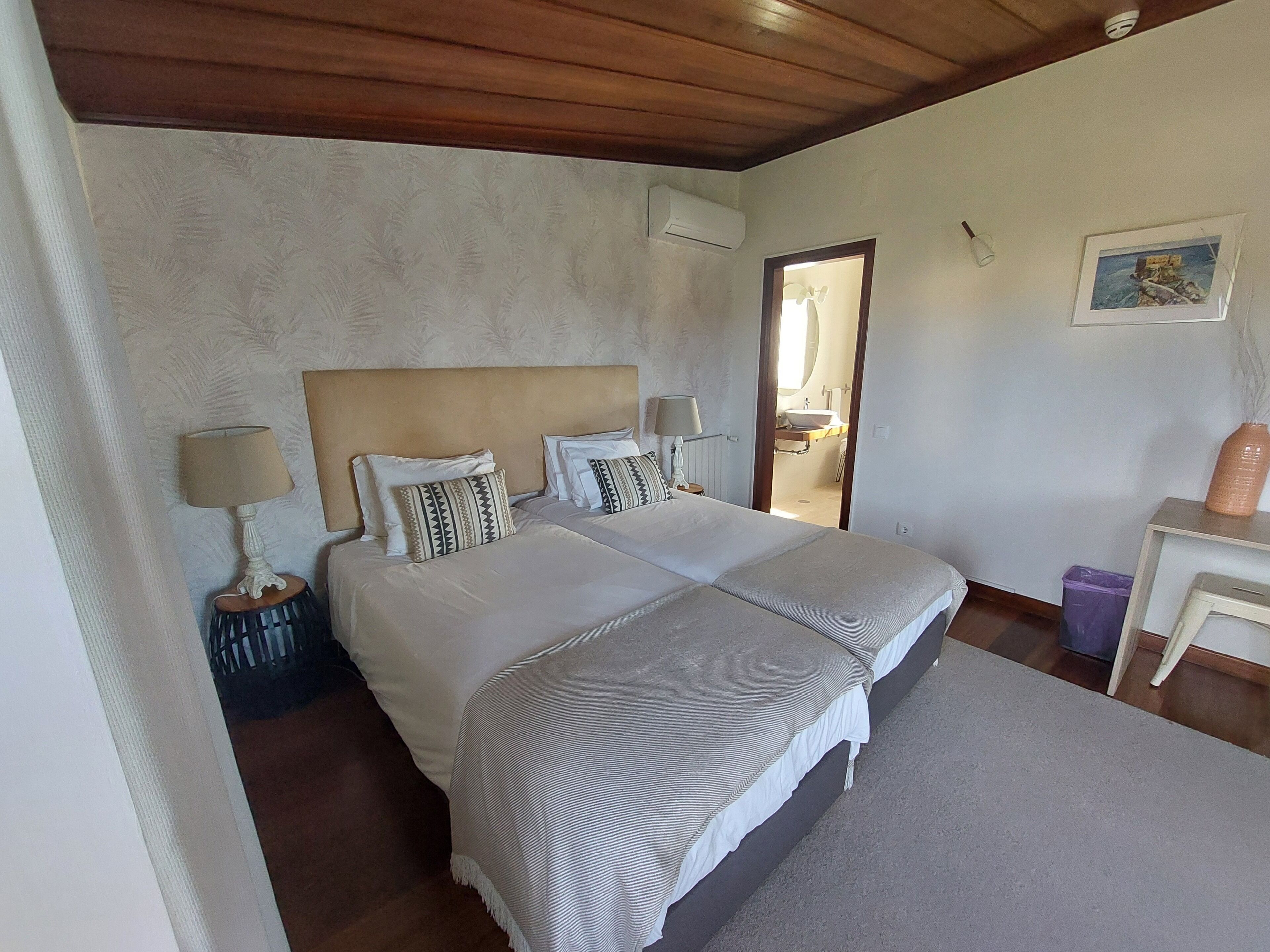 Un dormitorio con dos camas, techo de madera, pared blanca con estampado floral, mesita de noche con lámpara y una pequeña mesa con un jarrón.