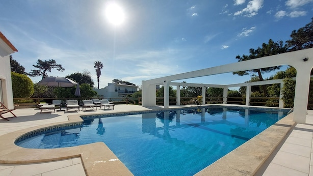 Una piscina con un pérgola blanca y sillones de descanso.