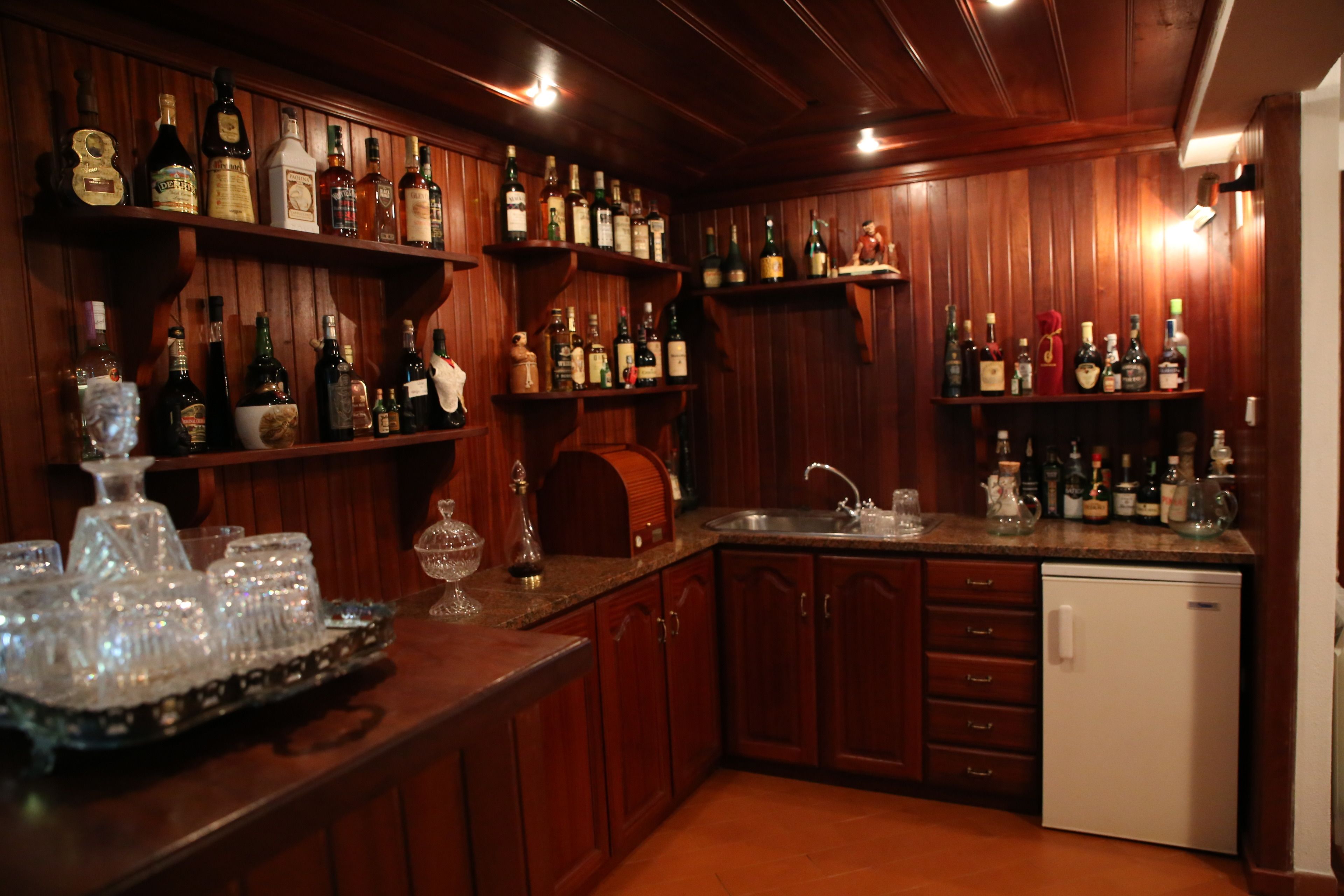 A bar area with wooden shelves stocked with bottles, glasses, and a refrigerator.