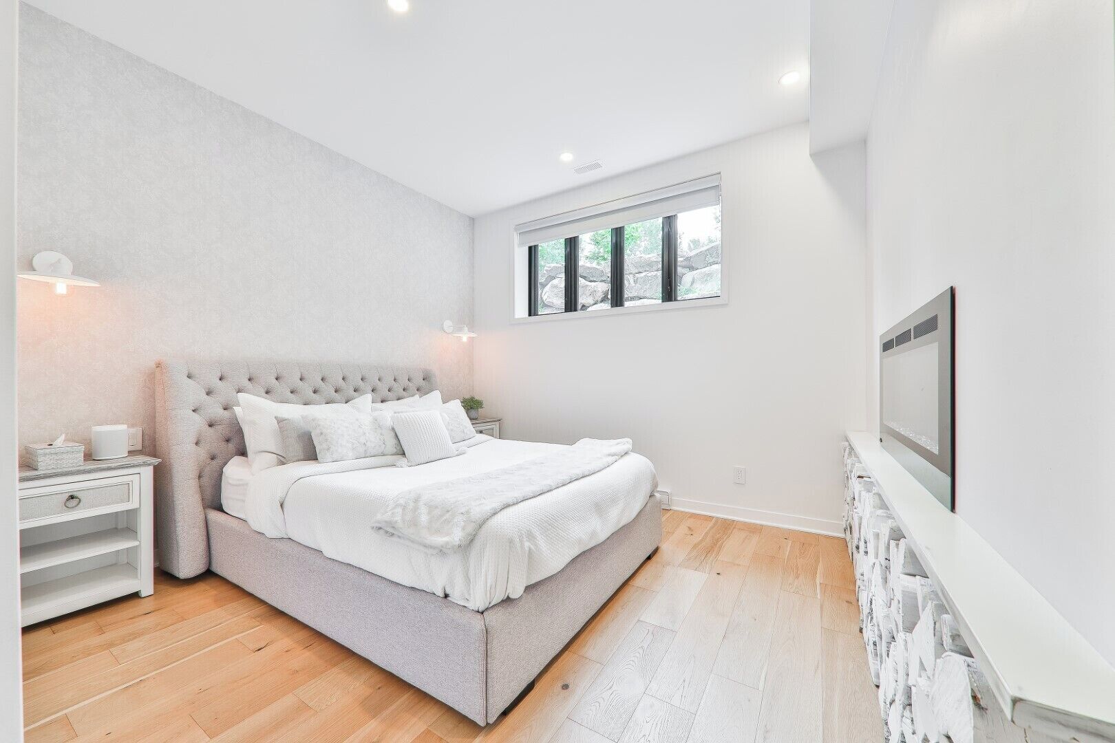 A modern bedroom with a large bed, a nightstand, a TV, and a window with a view of trees.