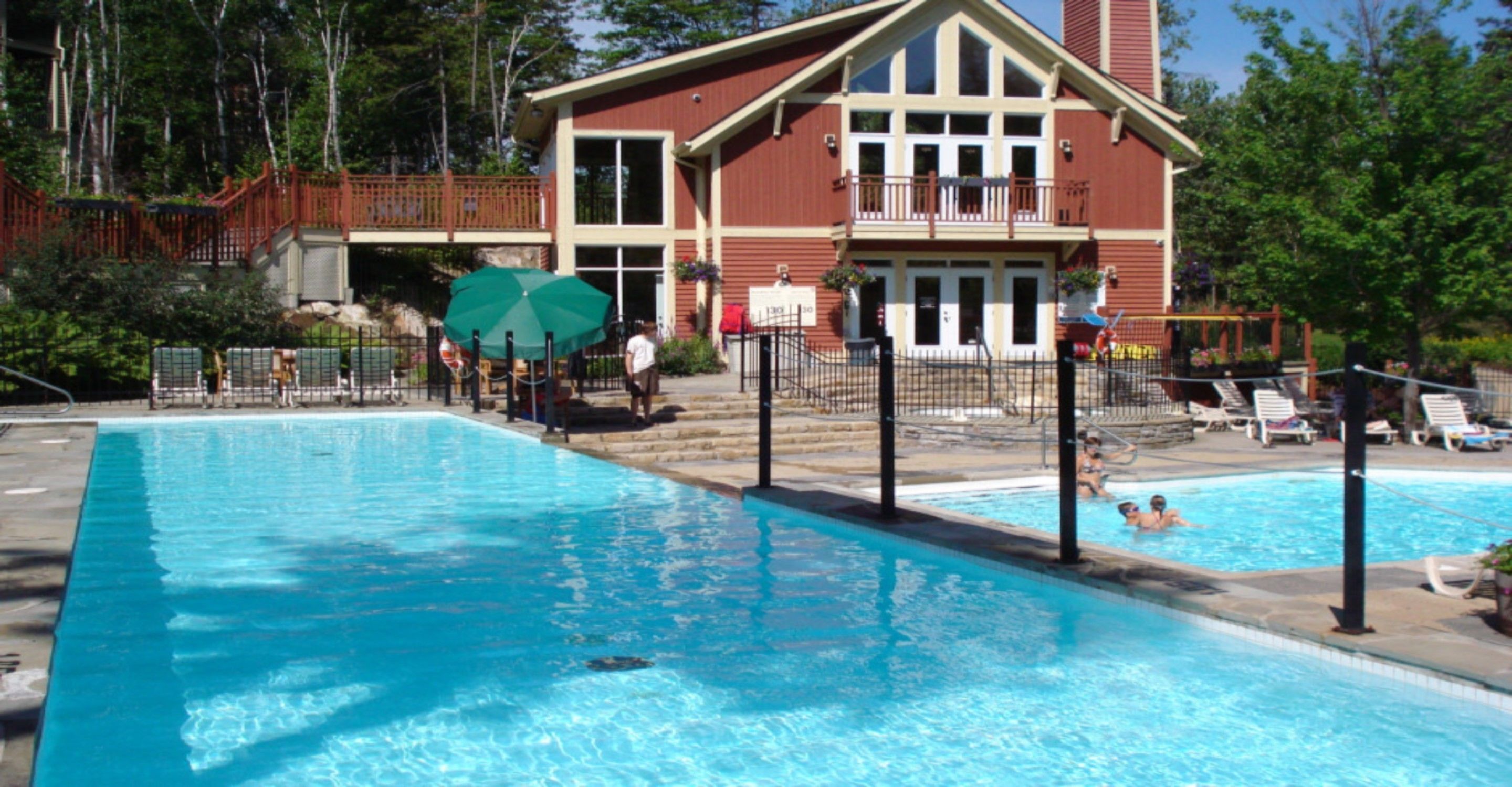 Una piscina con un edificio de madera al fondo, un área cercada y algunas personas en el agua.