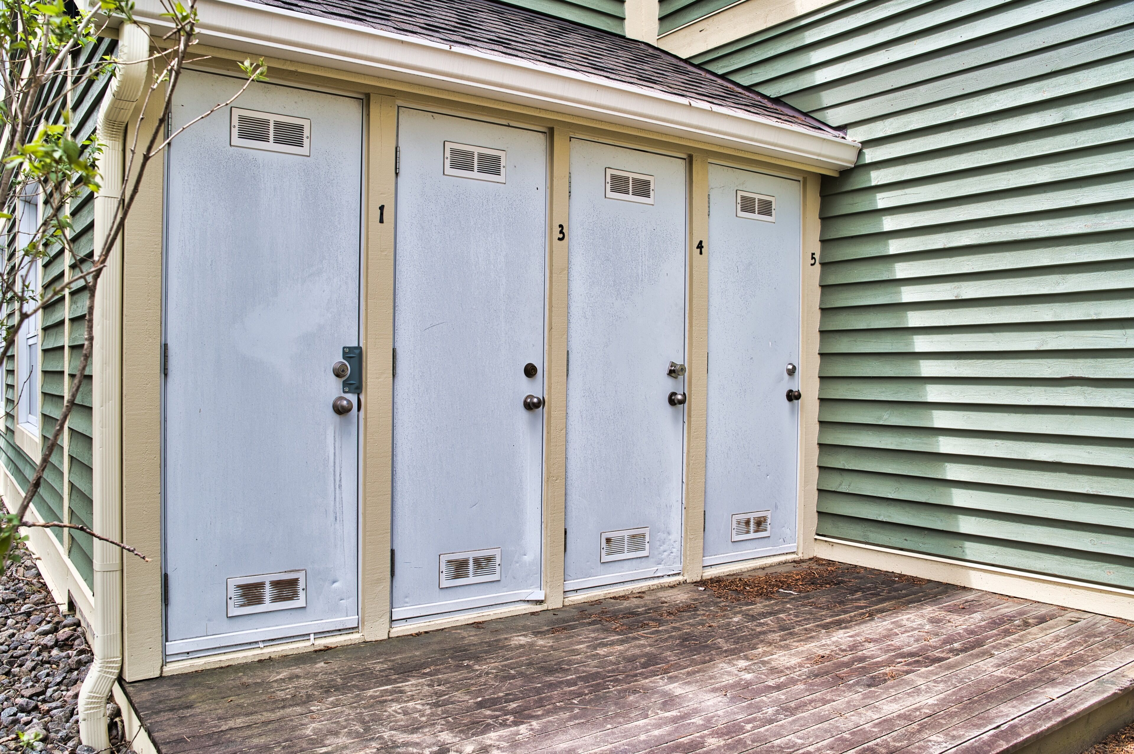 A row of four light blue doors with numbered vents, each with a door handle, against a green siding house.