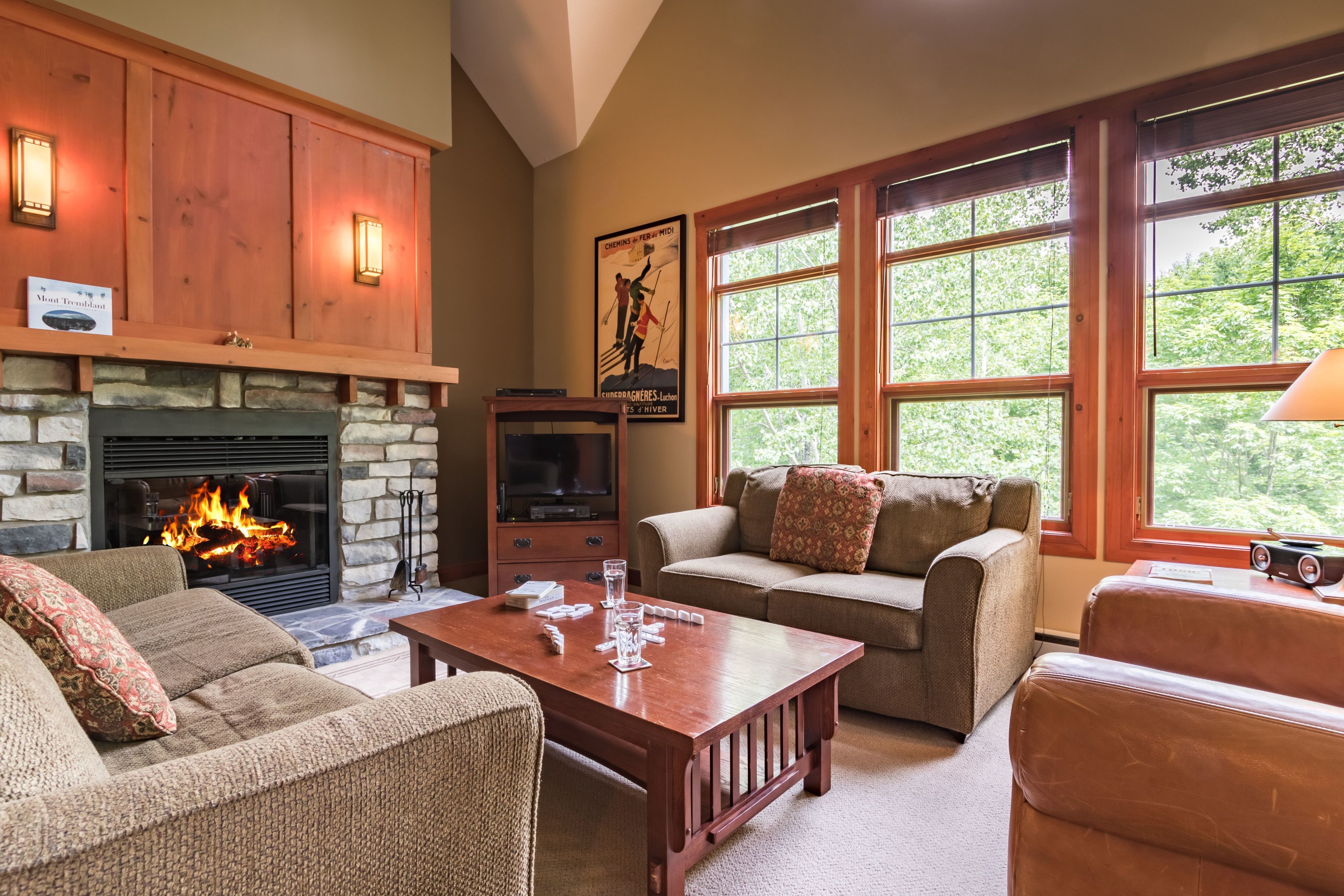 A cozy living room with a stone fireplace, wooden cabinets, and a large window offering a view of greenery.