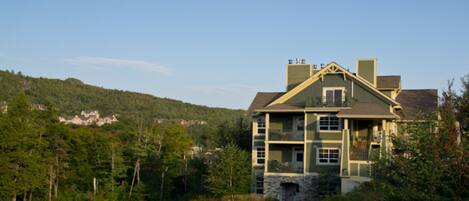 A two-story building with a stone foundation, multiple balconies, and a view of a forested hillside.