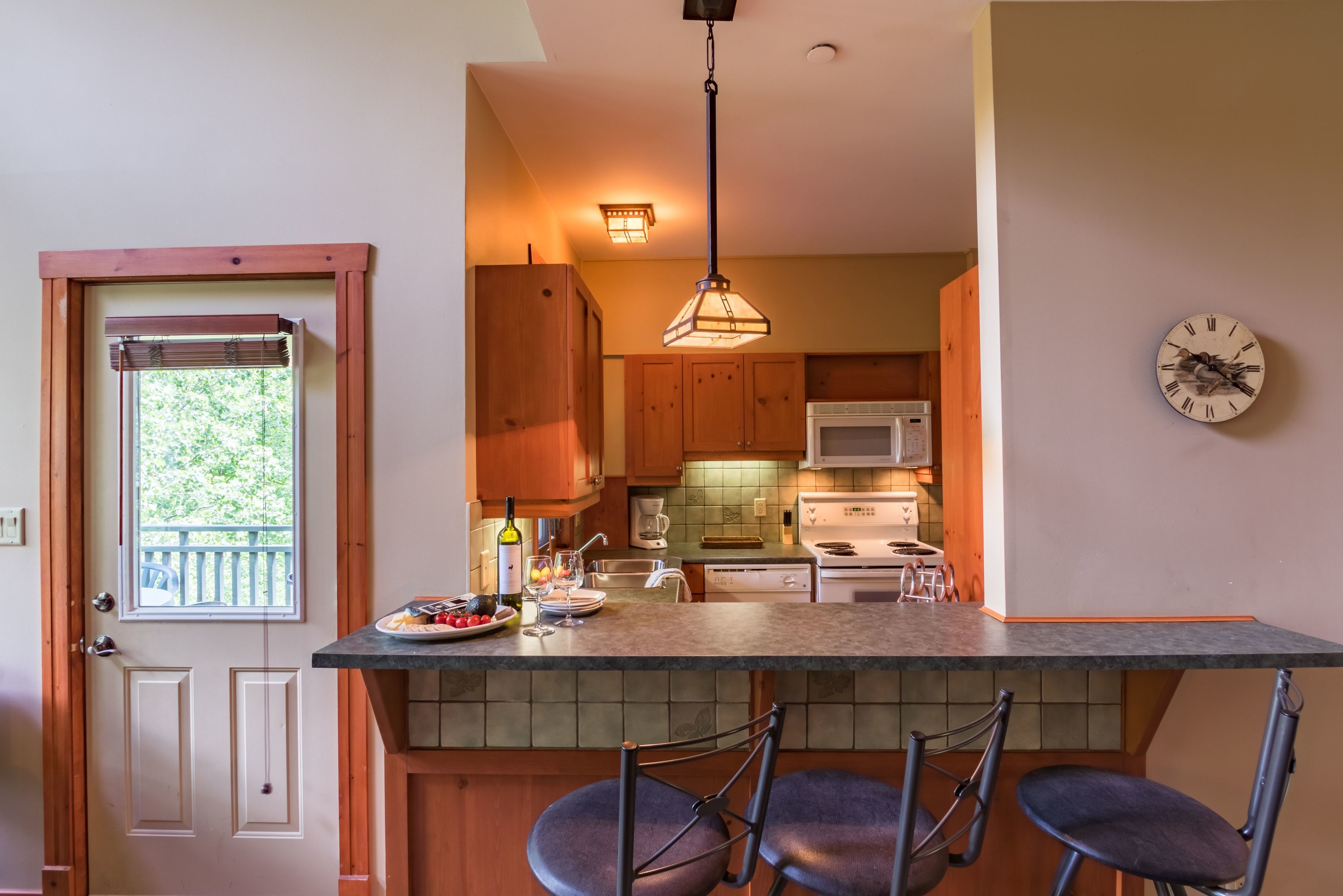 A kitchen with wooden cabinets, a microwave, a stove, a clock, and a bar area with chairs.
