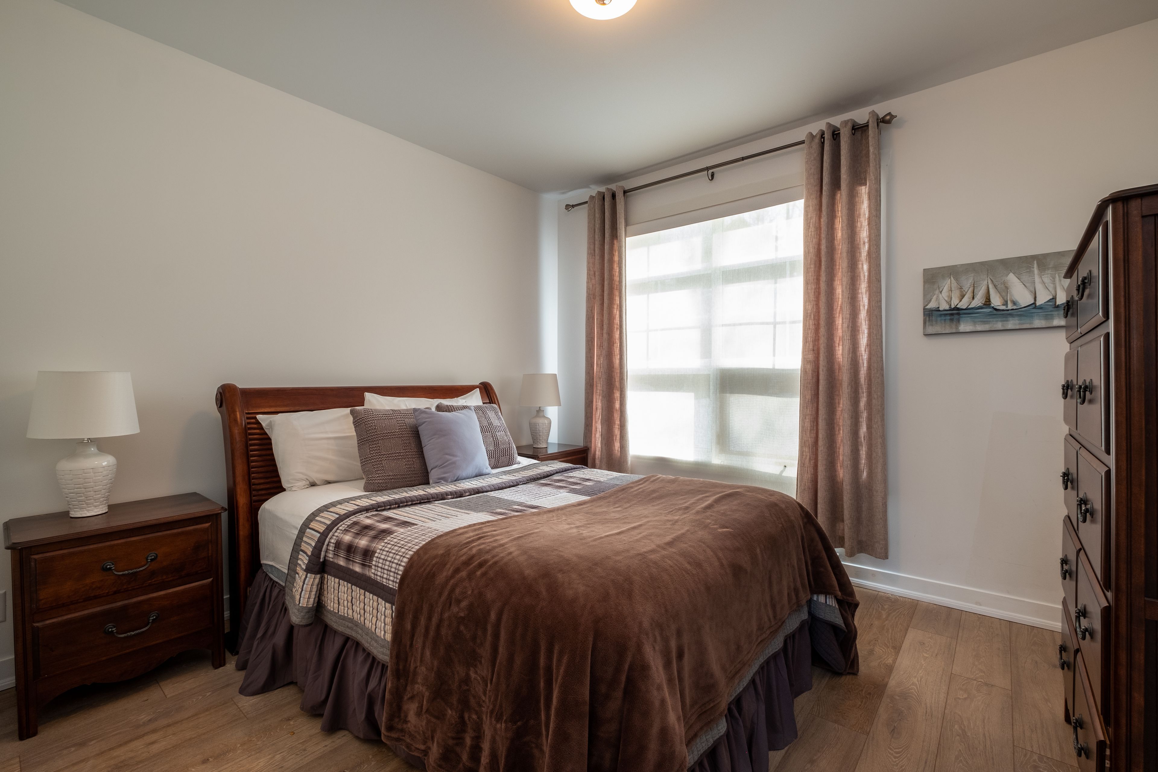 A bedroom with a wooden bed, a nightstand with a lamp, a dresser, and a window with curtains.