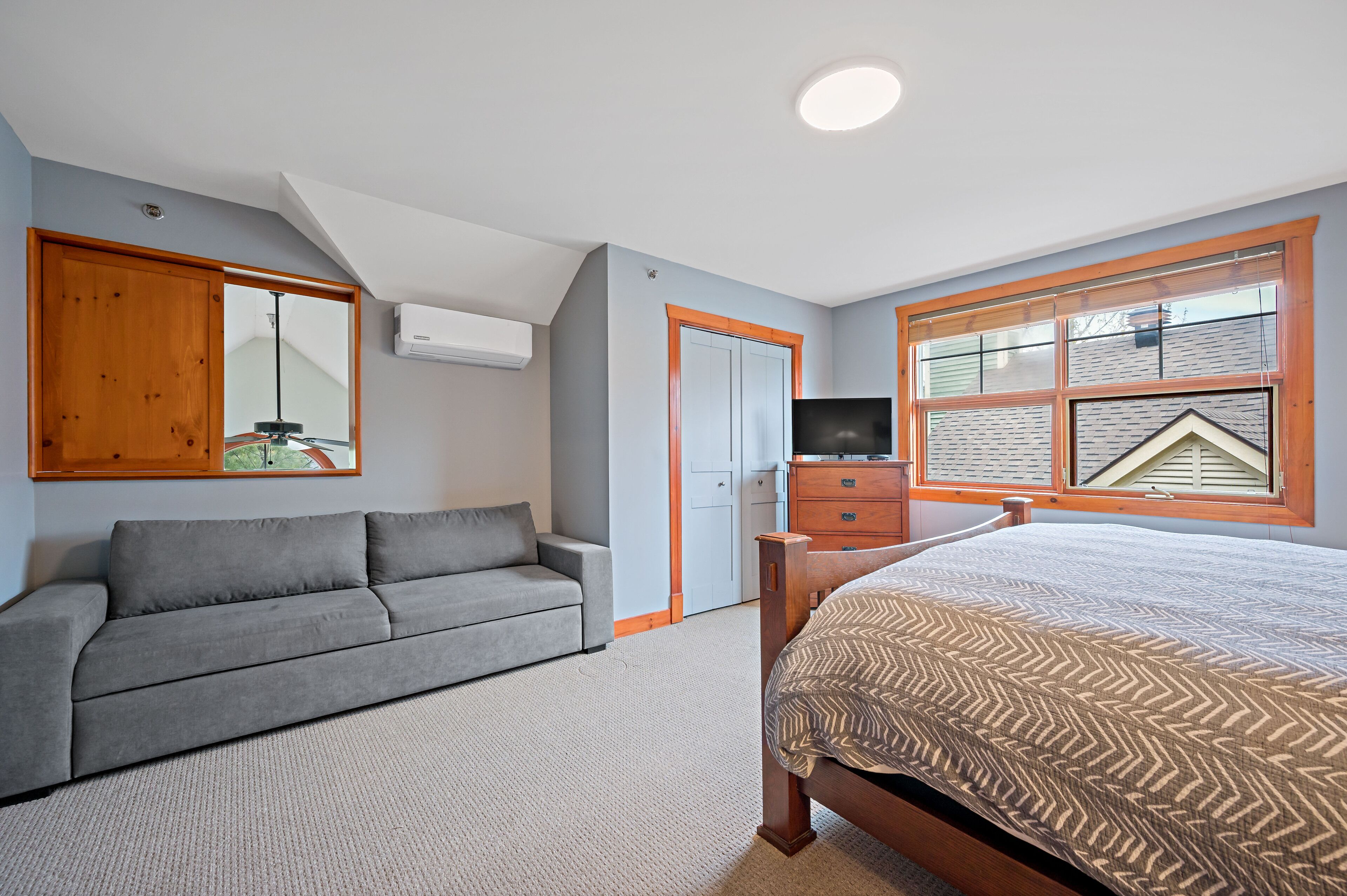 A bedroom with a bed, a gray sofa, a wooden wardrobe, and a window with blinds.