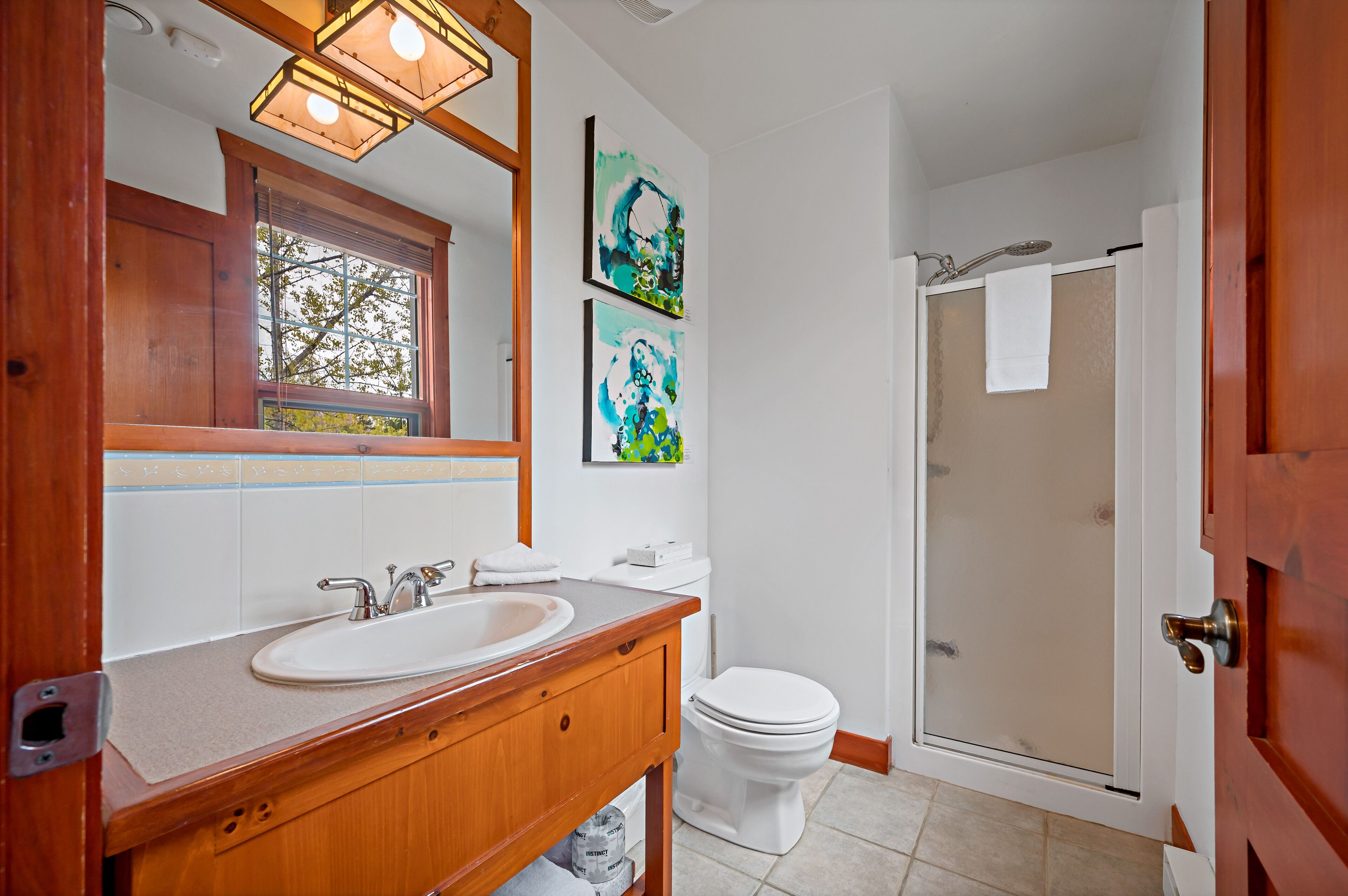A bathroom with a wooden vanity, white sink, and toilet. There are two framed artworks on the wall and a glass shower door.