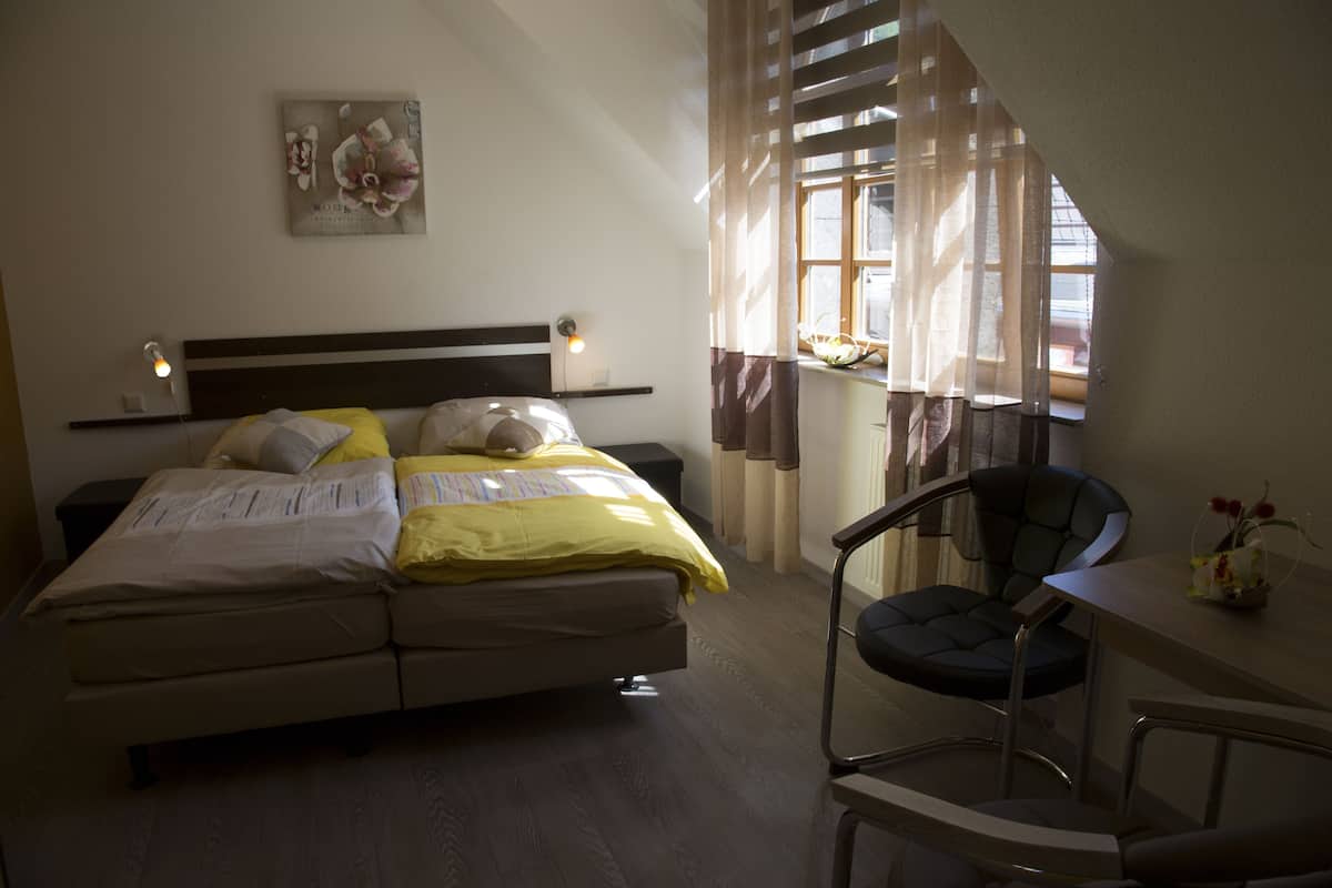 Een slaapkamer met een bed, nachtkastje, stoel en een raam met gordijnen.
