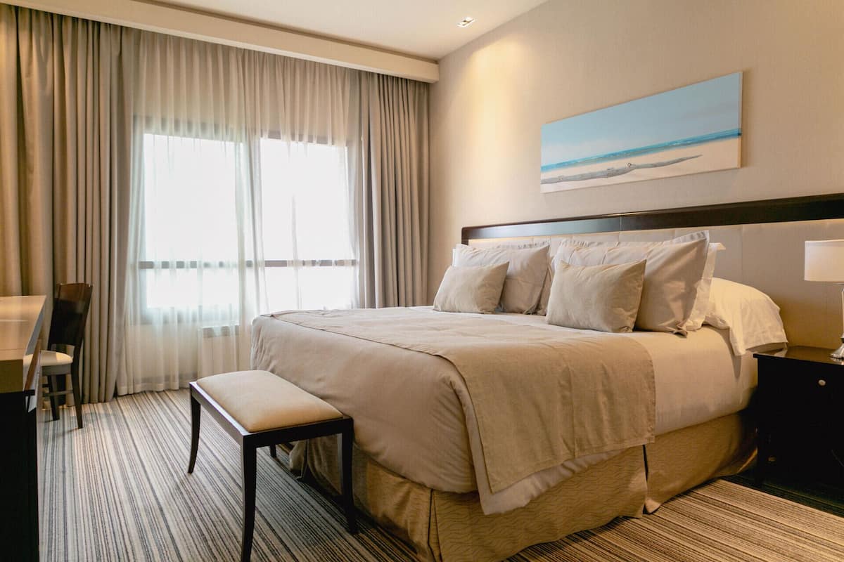 A hotel room with a large bed, a bench, a desk, and a painting of a beach scene.