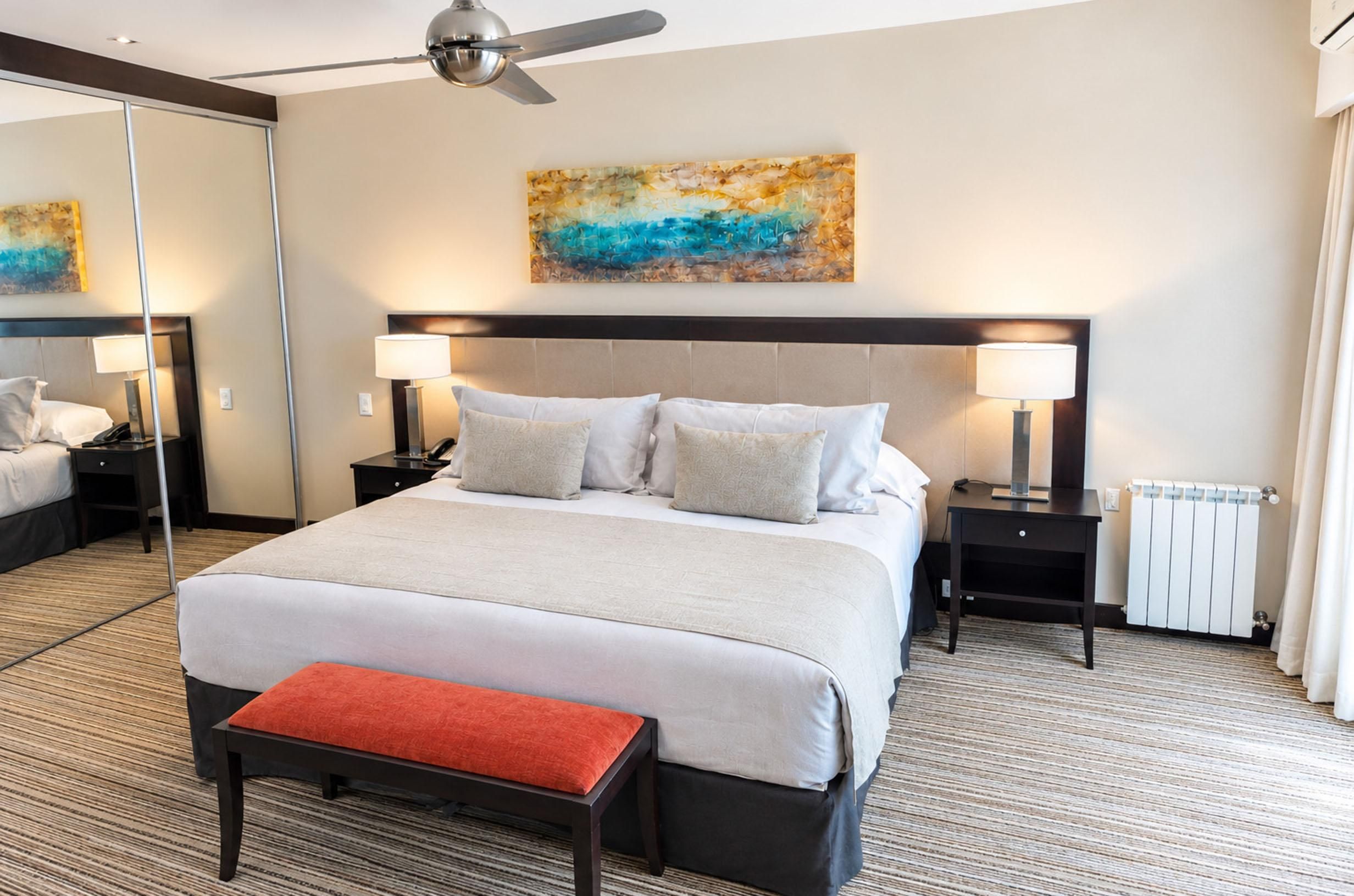 A hotel room with a large bed, two bedside tables, a floor lamp, a ceiling fan, a radiator, and a painting on the wall.