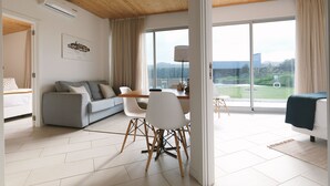 Una sala de estar moderna con sofá, mesa de comedor y sillas, ventanas amplias con vistas a zonas verdes y edificios, y un ventilador de techo.