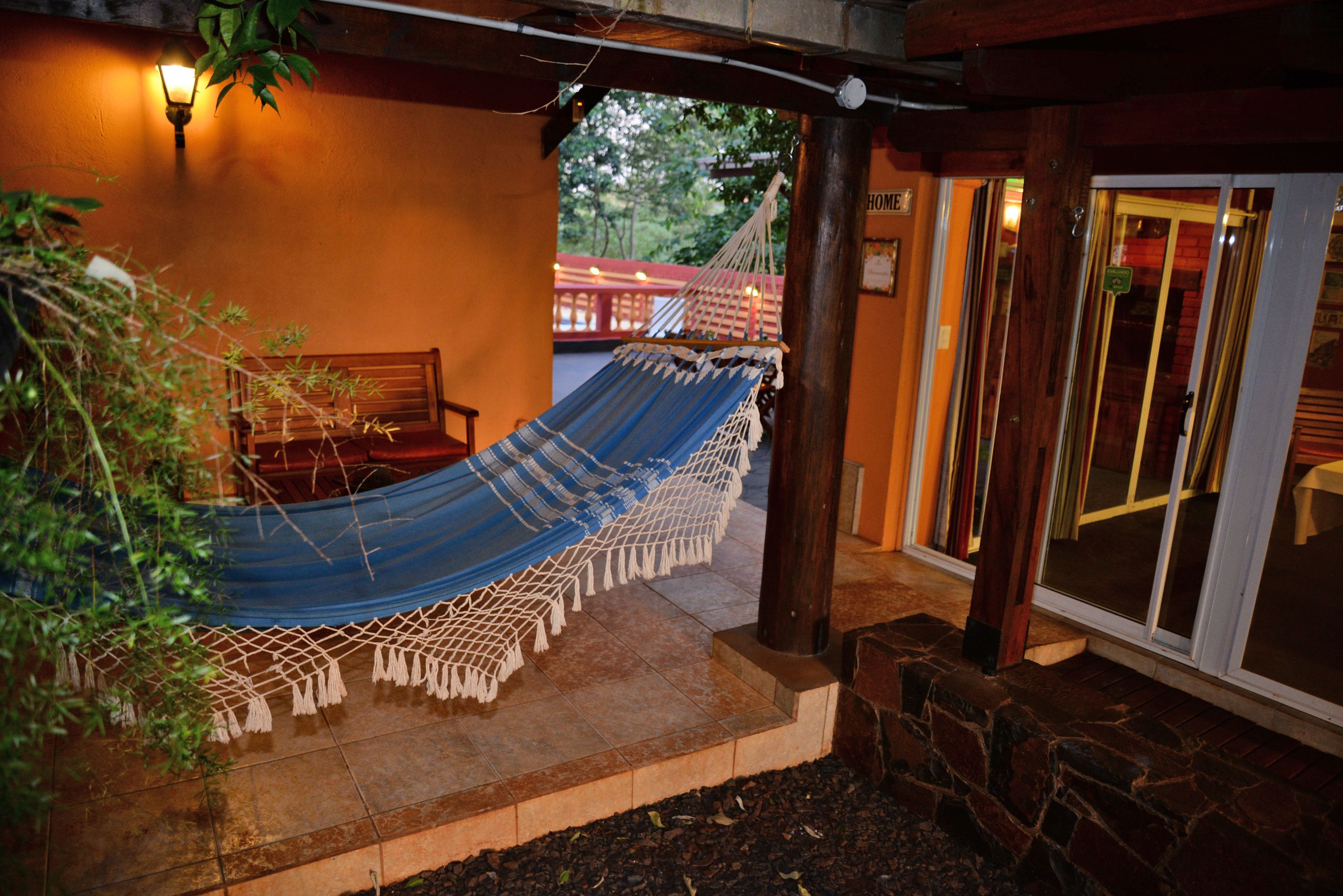 A hammock under a wooden pergola with a tiled patio, a bench, and a view of a red bridge in the background.