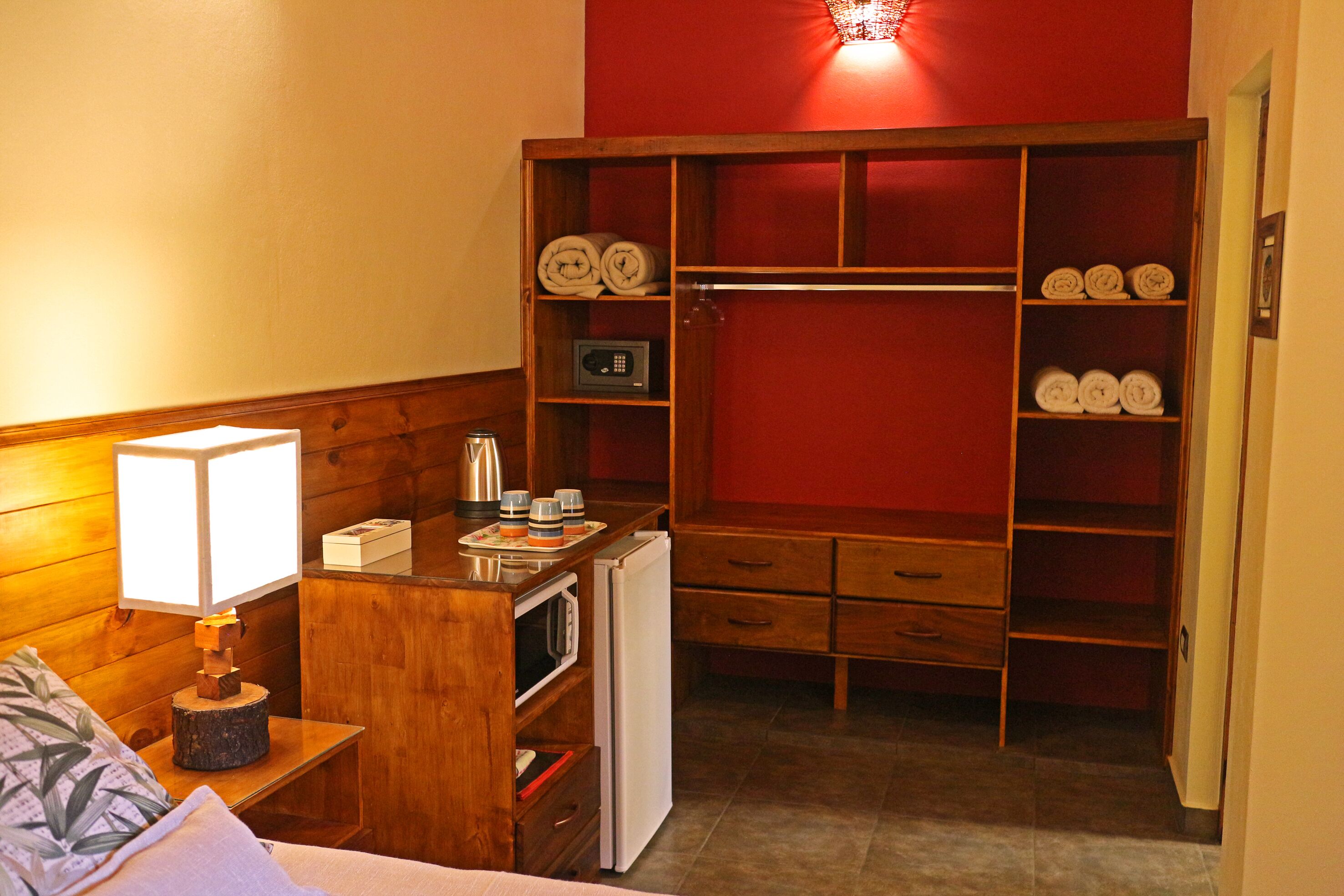 A compact hotel room with a wooden wardrobe, a small kitchenette, a bedside lamp, and a bed with a pillow.