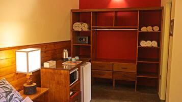 A compact hotel room with a wooden wardrobe, a small kitchenette, a bedside lamp, and a bed with a pillow.