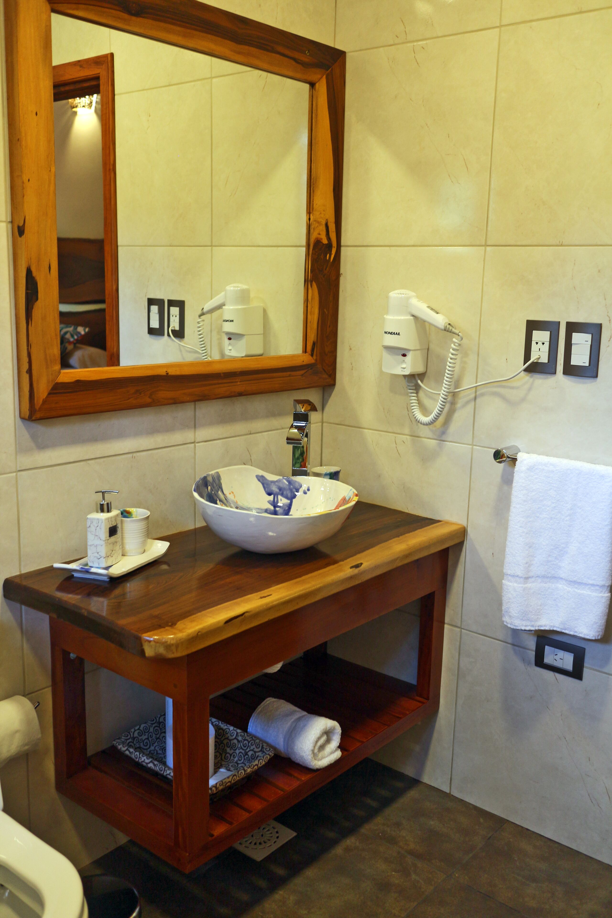 Casa de banho com toucador de madeira, um espelho grande, lavatório em cuba, secador de cabelo e barra de toalhas.