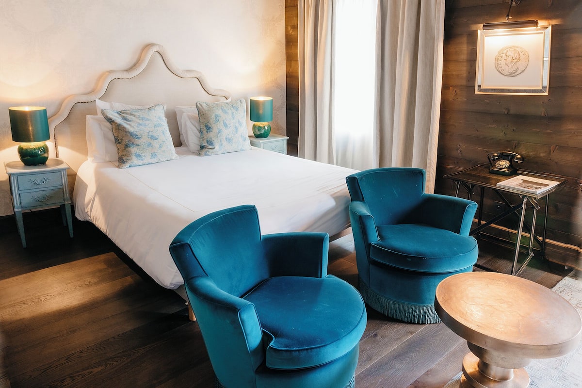 Ein Hotelzimmer mit einem großen Bett, zwei gleichartige Sessel, einem kleinen Tisch, einem Nachttisch mit Lampe und einem an der Wand montierten Telefon.