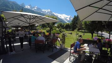 Un área de comedor al aire libre con mesas y sillas bajo grandes sombrillas, con montañas nevadas al fondo.