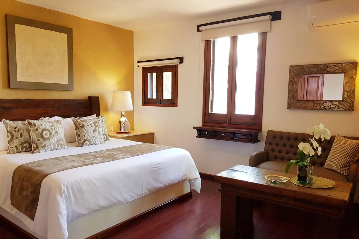 Une chambre d’hôtel avec un grand lit, une table de chevet en bois, une lampe de chevet, une fenêtre avec des rideaux, un canapé et un miroir décoratif.