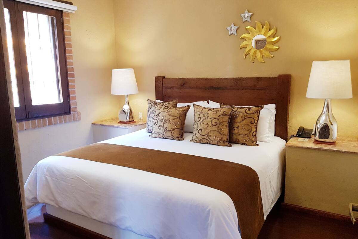 Un lit bien fait, avec une tête de lit marron, une literie blanche et des coussins décoratifs. On y trouve deux tables de chevet avec des lampes, et une décoration murale au-dessus du lit.