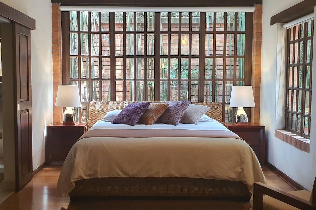 Une chambre à coucher bien rangée, avec une grande fenêtre, un lit agrémenté de plusieurs oreillers, deux tables de chevet avec des lampes et un banc en bois.