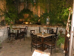 Un patio al aire libre con suelo de piedra, mesas y sillas de madera, y diversas plantas en macetas.