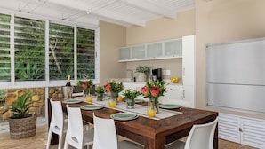 Un comedor con mesa de madera, sillas blancas, una cocina con armarios blancos y vistas a zonas verdes a través de ventanas amplias.