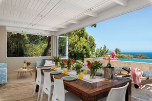 A dining area with a wooden table, white chairs, and a view of the ocean. - 15 Ottawa (Cape Town)