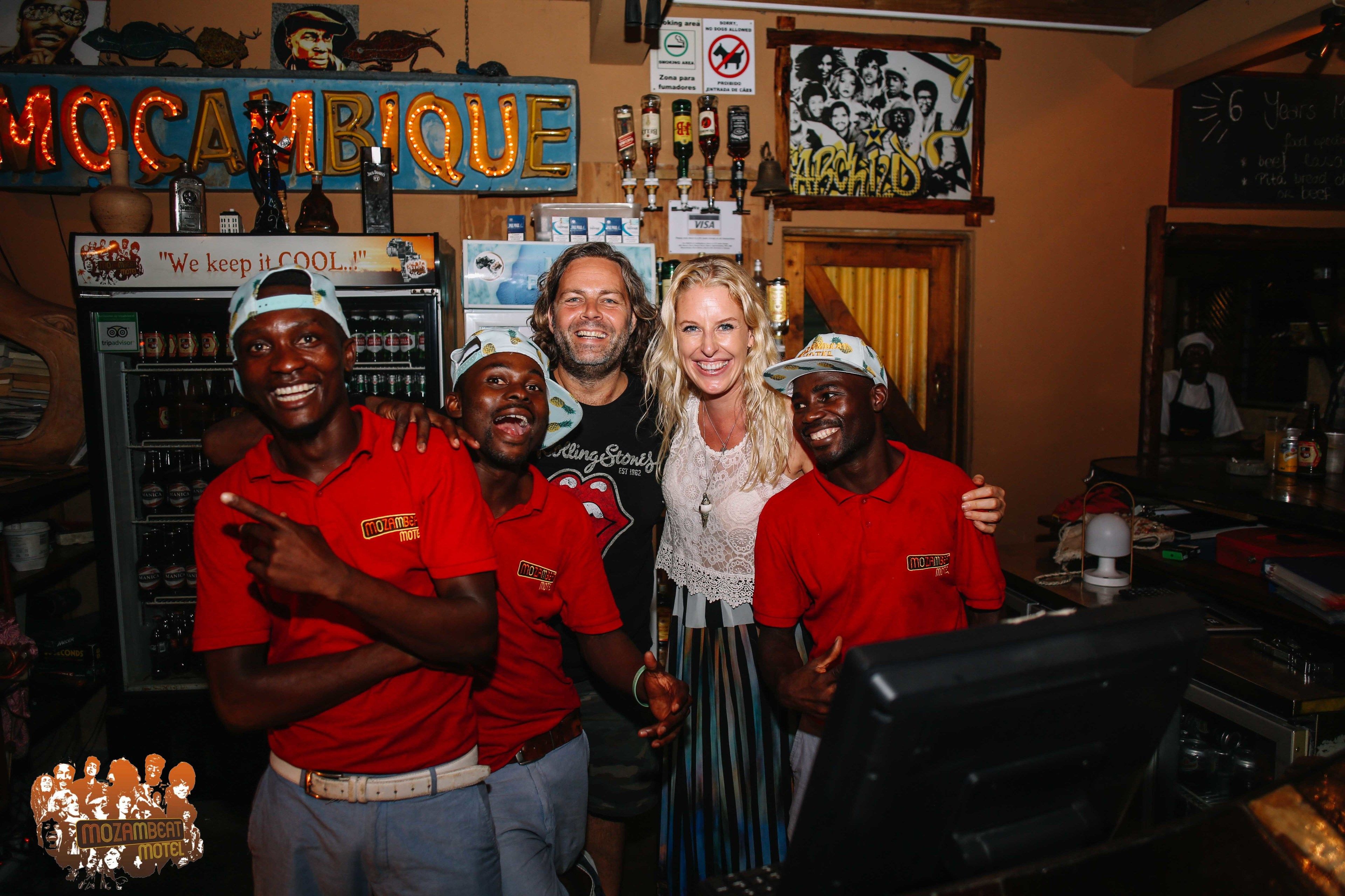 Ryhmä ihmisiä baarissa, taustalla kyltti, jossa lukee ”MOZAMBIQUE”, ja jääkaappi, joka on täynnä juomia.